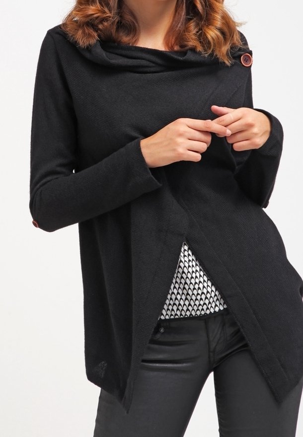 Femme portant un pull noir à manches longues avec des boutons, tenant le devant ouvert pour montrer une chemise à motif noir et blanc en dessous.