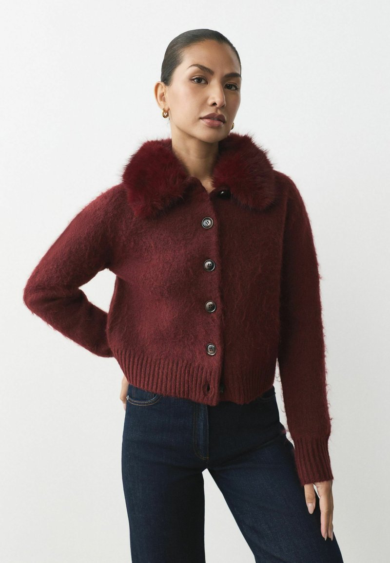 Bordeaux geknipte cardigan met een faux fur kraag, ronde knopen, geribbelde zoom en een gestructureerde stof. Gekleed met donkere denim jeans.