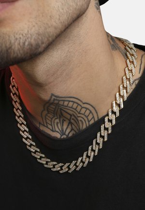 Collier en chaîne en ton doré avec des segments polis interconnectés ornés de pierres claires, reposant sur une surface de peau texturée et tatouée.