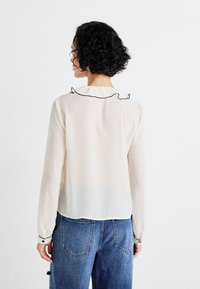 Light cream-colored blouse with a ruffled black collar, long sleeves, and button accents at the wrists. Worn with blue denim jeans.