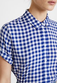 Chemise à manches courtes en vichy bleue et blanche avec un col boutonné et des boutons blancs qui descendent sur le devant.