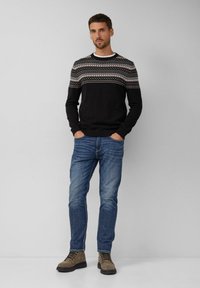 Mężczyzna stoi z rękami w kieszeniach, ubrany w wzorzysty czarny sweter, niebieskie dżinsy i brązowe sznurowane buty na tle jednolitego szarego tła.