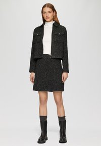 Schwarze strukturierte, cropped Jacke mit zwei Fronttaschen, kombiniert mit einem figurbetonten schwarzen Rock. Schwarze Stiefeletten mit dicker Sohle und grauen Socken.