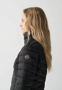 Zwarte pufferjack met een hoge kraag en gewatteerd design. Logo patch op de linkerarm. Gladde textuur, slim fit.