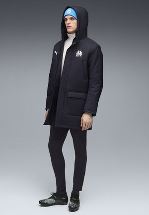 Male model wearing a black hooded Puma jacket with Olympique de Marseille logo, blue beanie, black leggings, and black cleats, standing against gray.