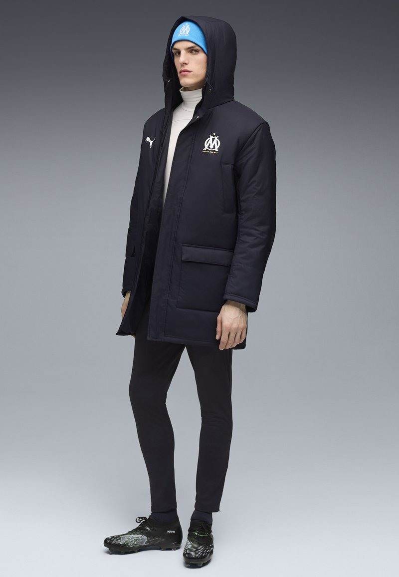 Modèle masculin portant une veste noire à capuche Puma avec le logo de l’Olympique de Marseille, un bonnet bleu, des leggings noirs et des crampons noirs, debout sur fond gris.