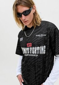 Jeune personne avec de longs cheveux portant des lunettes de soleil noires, un t-shirt noir de l'UFC (Ultimate Fighting Championship) par-dessus des manches longues blanches, une chaîne en argent et des boucles d'oreilles en or.