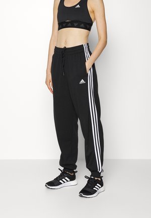 Vrouw draagt een zwarte Adidas sportbeha, zwarte joggingbroek met witte strepen en zwarte Adidas sneakers, staande tegen een witte muur.