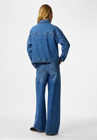 Femeie cu spatele spre cameră, purtând o jachetă denim albastră și blugi evazați, pe fundal alb simplu.