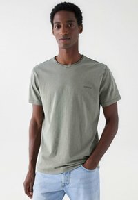 Olivově zelené bavlněné tričko s kulatým výstřihem, krátkými rukávy a diskrétním logem značky na hrudi. Spárováno se světle modrými džíny.