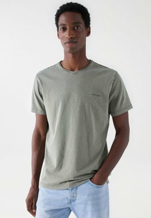 Camiseta de algodón verde oliva con cuello redondo, mangas cortas y un sutil logo de la marca en el pecho. Combinada con jeans de color azul claro.