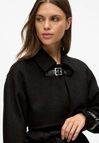 Czarny wełniany płaszcz z załamanym kołnierzem, z czarnym skórzanym zapięciem i pasującymi akcentami. Projekt obejmuje długie rękawy i zapięcie w talii.