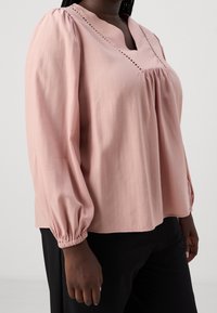 Blusa rosa chiaro con scollo a V, maniche a sbuffo e cuciture decorative lungo il collo. Tessuto liscio con sottili righe verticali.
