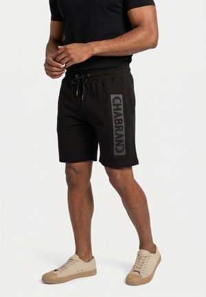 Man in zwarte short met trekkoord en tekst "CHABRAND" op de linker dij, zwart shirt en beige sneakers, staand tegen een effen achtergrond.