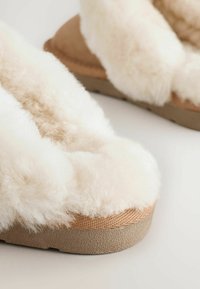 Pehmeät valkoiset tossut, joissa on beigenvärinen pohja, pehmeä ja pörröinen yläosa sekä teksturoitu, pitoa parantava ulkopohja.