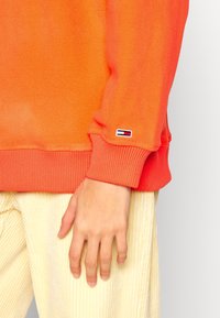 Zbliżenie na dłoń opierającą się z boku, ubraną w pomarańczową polarową rękawicę z małym prostokątnym logo w kolorach czerwonym, białym i niebieskim oraz w beżowe sztruksowe spodnie.