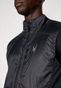 Melnā izolētā veste ar augstu apkakli, ar rāvējslēdzēju priekšpusē un ar zirneklis logotipu uz krūtīm. Gluda, spīdīga auduma ar vieglu dizainu.