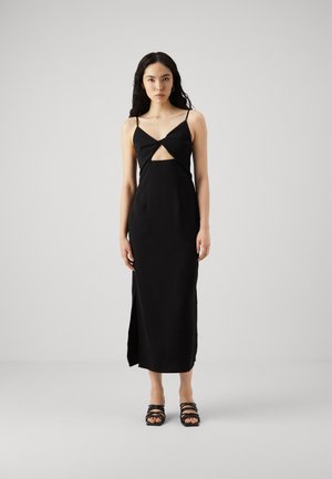 Black sleeveless dress with a twist front design. Smooth fabric, mid-length, side slit, paired with strappy black sandals.