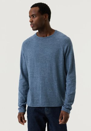 Man draagt een blauw overhemd met lange mouwen en donkere spijkerbroek, kijkt naar links tegen een effen lichte achtergrond.