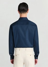 Marineblaues Langarmhemd mit glatter Textur und spitzem Kragen, kombiniert mit cremefarbenen Hosen mit Gesäßtaschen und goldfarbenen Akzenten.