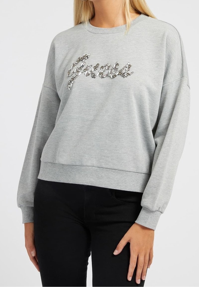 Femme portant un sweat-shirt gris clair avec le mot « Greece » en sequins argentés sur la poitrine, associé à un pantalon noir.