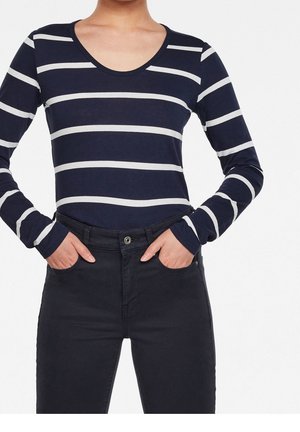 Kobieta w granatowo-białej paskowanej koszuli z długim rękawem, wpuszczonej w wysokowalne czarne spodnie, z rękami w kieszeniach.