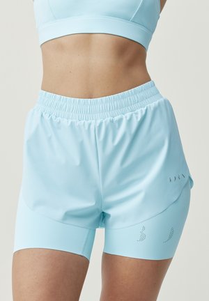 Utrzymana w dobrej formie kobieta w jasnoniebieskich warstwowych sportowych szortach i dopasowanym sportowym staniku, pokazana od połowy tułowia do połowy uda.