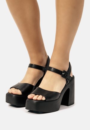 Sandales plates noires avec une large bride avant, une bride de cheville et un détails de boucle. Matériau en cuir lisse avec un design à talon épais.