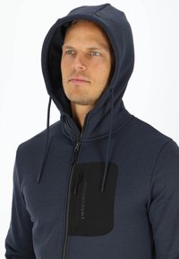 Felpa blu navy con full zip, cappuccio con cordino e una tasca sul petto con zip nera che presenta il testo "SWEDEMOUNT". Tessuto morbido e testurizzato.