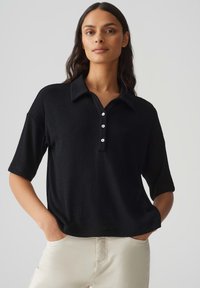 Polo shirt noir à manches courtes avec un col, doté d'une patte de boutonnage et d'une texture douce. Porté avec un pantalon de couleur claire.