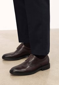 Scarpe da uomo in pelle marrone con punta arrotondata, dettagli di cucitura discreti e suola in gomma nera, abbinate a pantaloni scuri.