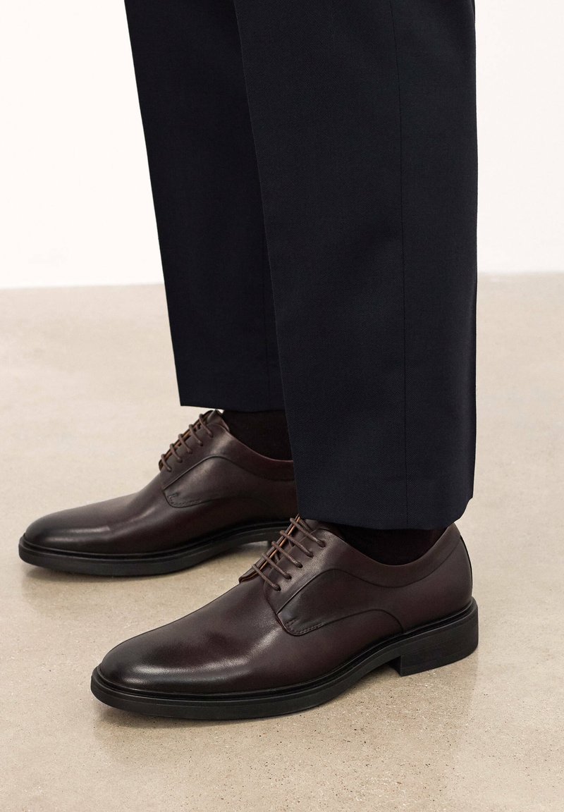 Scarpe da uomo in pelle marrone con punta arrotondata, dettagli di cucitura discreti e suola in gomma nera, abbinate a pantaloni scuri.
