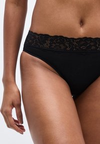 Μαύρο μπικίνι με δαντέλα στο τελείωμα, κατασκευασμένο από απαλό, ελαστικό ύφασμα και με σχεδιασμό χαμηλής μέσης. Η δαντέλα στη μέση με κυματιστά τελειώματα προσθέτει λεπτομέρεια.