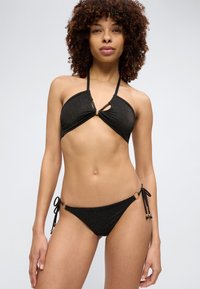 Γυναίκα με σγουρά μαλλιά, φορώντας μαύρο μπικίνι λαιμόκοψης τύπου halter και κάτω μέρος μπικίνι με δεσίματα στο πλάι, πάνω σε απλό φόντο.