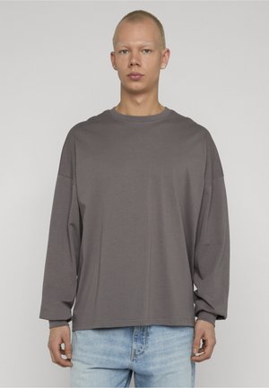 Młoda osoba z krótkimi włosami, ubrana w luźną szarą bluzkę z długim rękawem i jasnoniebieskie dżinsy, stojąca na jednolitym tle.