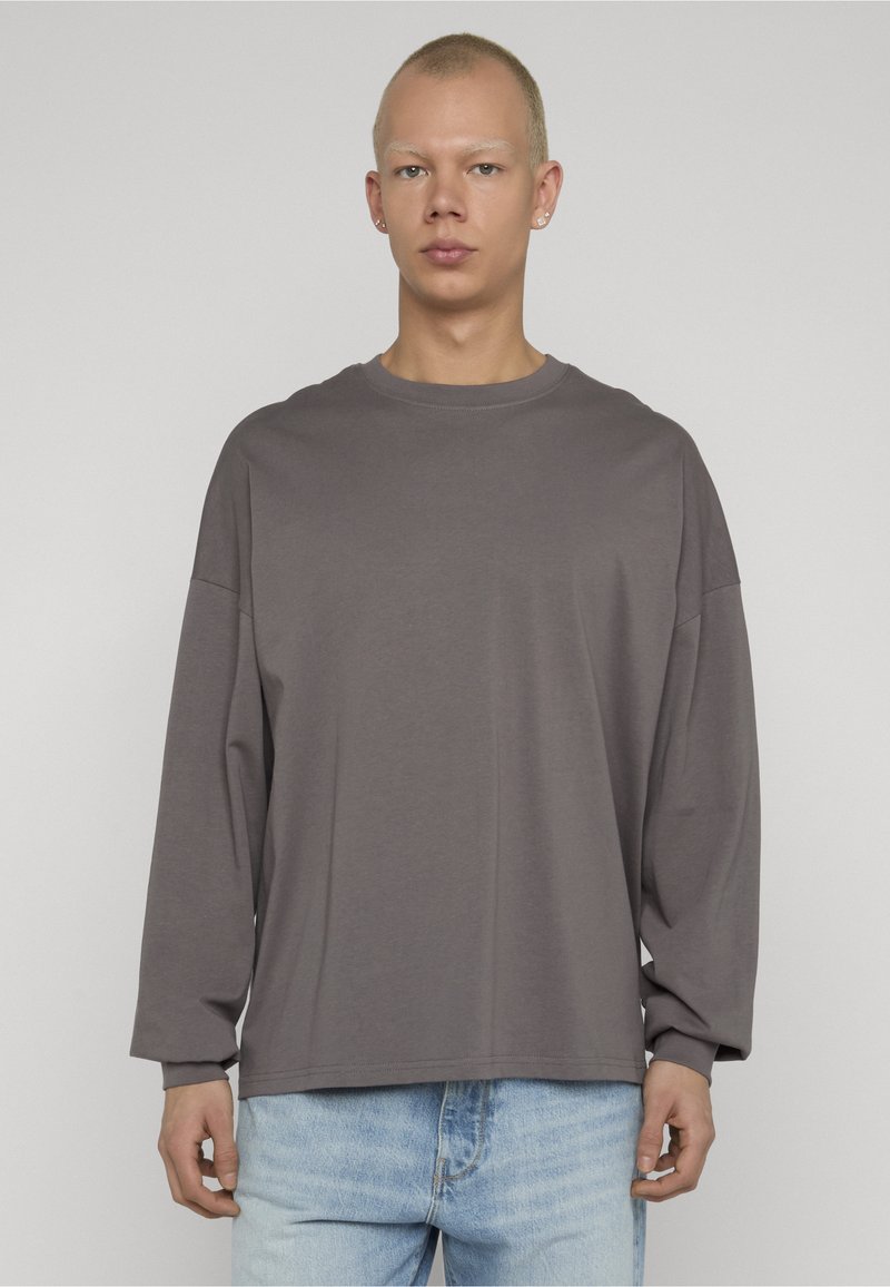 Young person with short hair wearing a loose gray long-sleeve shirt and light blue jeans, standing against a plain background.