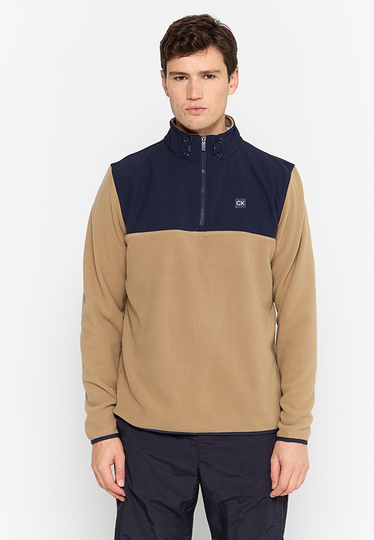 Pull en polaire beige avec un haut à zip quart bleu marine. Présente un logo CK sur la poitrine et une texture douce. Convient pour une tenue décontractée.