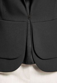 Schwarzer taillierter Blazer aus glattem Stoff, mit runden Revers und einem verdeckten Reißverschluss auf der Vorderseite. Texturierter Innenfutter sichtbar.