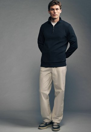 Navy zip-up pullover with textured fabric, paired with light beige pants. Shoes are gray with cream accents and a white sole.