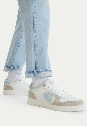 Nohy v bílých teniskách s světle modrými a béžovými detaily, doplněné bílými ponožkami a svinutými světle modrými džínami.