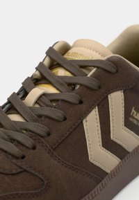 Sneaker in suede marrone con accenti beige, caratterizzato da una punta rotonda, chiusura con lacci e logo. Motivo a chevron distintivo sul lato.
