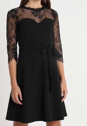 Kvinde iført en sort knælængde kjole med gennemsigtige blonder på trekvartærmer, blondeindlæg og et bindebælte i taljen.