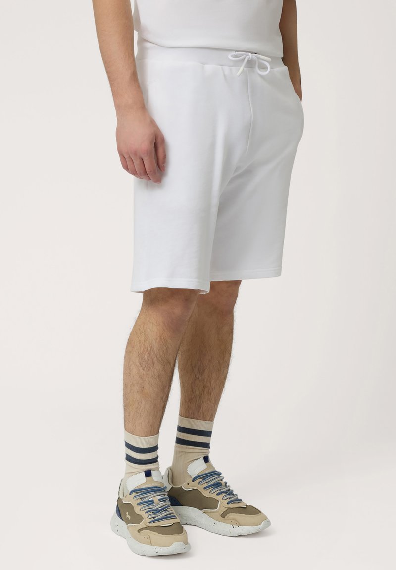 Shorts en coton blancs avec une taille élastique et un cordon de serrage. Portés avec des baskets beiges et des chaussettes rayées. Texture lisse, coupe décontractée.