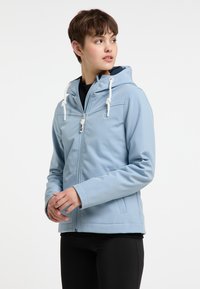 Šviesiai mėlynas zip-up tipo džemperis su kapuce, su raišteliais, lygia tekstūra ir šoninėmis kišenėmis. Dėvimas ant juodų drabužių.