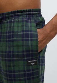 Grønne og marineblå ternede pyjamasshorts med elastisk talje og sidelomme. Har et sort mærke med hvid tekst på siden.