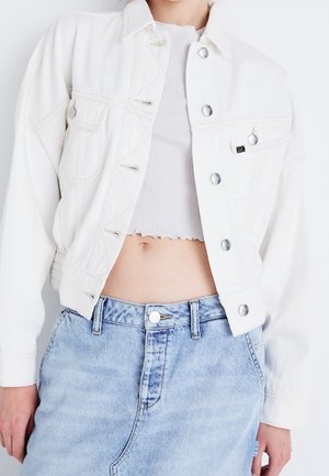 Personne portant une veste courte blanche par-dessus un top court côtelé blanc et un jean en denim bleu clair taille haute.