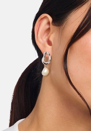 Unregelmäßiger silberner Creolen-Ohrring mit einem einzelnen weißen Perltropfen, getragen am rechten Ohr einer Frau mit zurückgebundenem dunkelbraunem Haar.