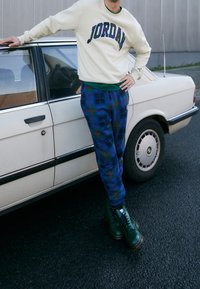 Felpa cremisi con scritta "JORDAN", maglietta verde, pantaloni della tuta a quadri blu e verdi e stivali verdi, appoggiata a un'auto beige.