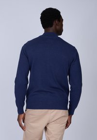 Pull en maille bleu marine, manches longues, col haut, texture lisse, design ajusté, affiche un style minimaliste sans logos visibles.
