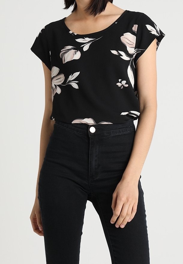 Femme portant un haut noir à manches courtes fleuri, rentré dans un jean noir taille haute, sur un fond uni.